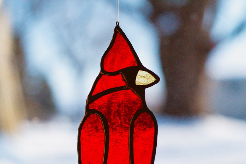 Op de afbeelding: Een rood glas-in-lood ornament van een kardinaal dat aan een raam hangt. De kardinaal is gedetailleerd met zwarte contouren en een gele snavel.