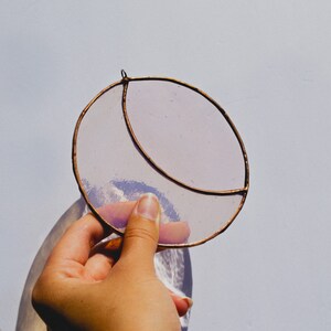 May include: A hand holds a stained glass crescent moon suncatcher. The suncatcher is made of clear glass with a copper wire frame. The glass has a light purple tint.