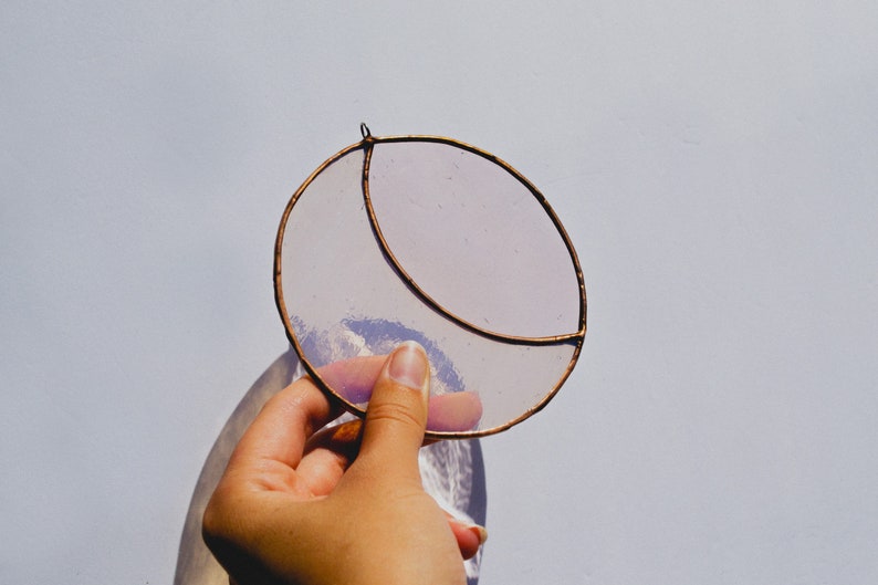 May include: A hand holds a stained glass crescent moon suncatcher. The suncatcher is made of clear glass with a copper wire frame. The glass has a light purple tint.