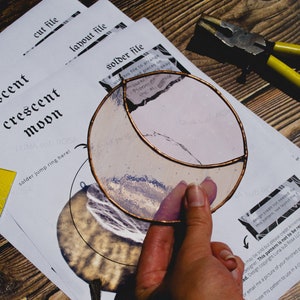 May include: A stained glass crescent moon with a copper frame. The moon is a light pink color with a white and blue design. The image also shows a pair of pliers and a sheet of paper with the words "crescent moon" printed on it.