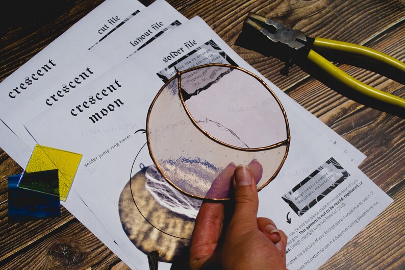 May include: A stained glass crescent moon with a copper frame. The moon is a light pink color with a white and blue design. The image also shows a pair of pliers and a sheet of paper with the words "crescent moon" printed on it.