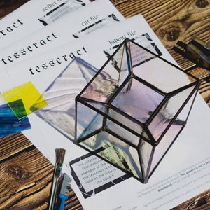K&ouml;nnte beinhalten: Ein klarer Glasw&uuml;rfel mit schwarzem Metallrahmen, der auf einer Holzoberfl&auml;che liegt. Der W&uuml;rfel ist ein Tesserakt, ein vierdimensionales Analogon des W&uuml;rfels. Der Text "Der Tesserakt ist zum W&uuml;rfel, wie der W&uuml;rfel zum Quadrat ist" ist auf einem Blatt Papier sichtbar.