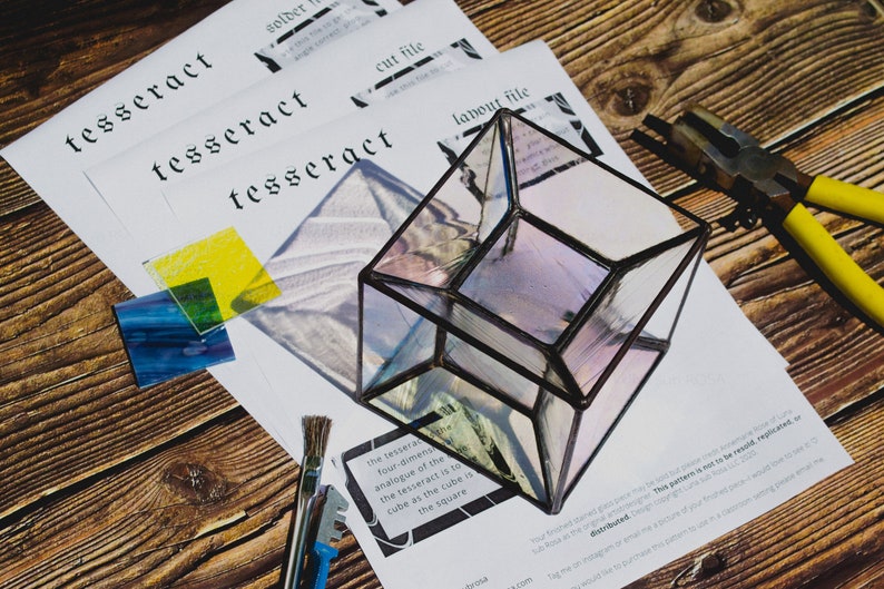 K&ouml;nnte beinhalten: Ein klarer Glasw&uuml;rfel mit schwarzem Metallrahmen, der auf einer Holzoberfl&auml;che liegt. Der W&uuml;rfel ist ein Tesserakt, ein vierdimensionales Analogon des W&uuml;rfels. Der Text "Der Tesserakt ist zum W&uuml;rfel, wie der W&uuml;rfel zum Quadrat ist" ist auf einem Blatt Papier sichtbar.
