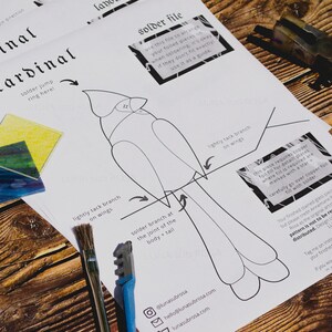 Op de afbeelding: Een zwart-witte lijntekening van een kardinaal op een wit vel papier. Het papier bevat instructies voor het solderen van gekleurde glasstukken om een kardinaal te maken. De instructies omvatten "solder jump ring here!" en "lightly tack branch on wings".