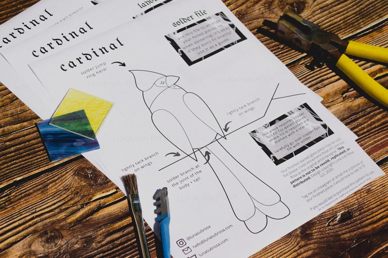 Op de afbeelding: Een zwart-witte lijntekening van een kardinaal op een wit vel papier. Het papier bevat instructies voor het solderen van gekleurde glasstukken om een kardinaal te maken. De instructies omvatten "solder jump ring here!" en "lightly tack branch on wings".