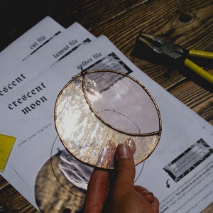 May include: A stained glass crescent moon with a copper foil border. The moon is made of clear glass with a silver glitter effect. The moon is being held by a hand. The image also includes instructions for making a stained glass crescent moon.