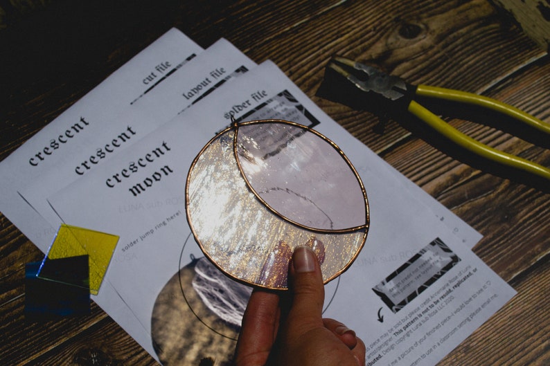 May include: A stained glass crescent moon with a copper foil border. The moon is made of clear glass with a silver glitter effect. The moon is being held by a hand. The image also includes instructions for making a stained glass crescent moon.