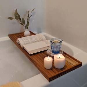 May include: A wooden bath tray with a book, two candles, and a glass of water with a lemon slice. The tray is in a bathtub with bubbles.