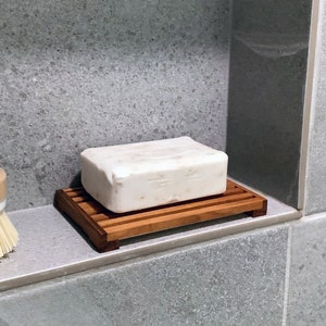 May include: A white bar of soap sits on a wooden soap dish. The soap dish is made of light brown wood with a slatted design. The soap dish is sitting on a grey tiled surface.