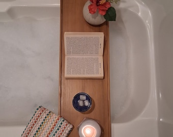 Puede incluir: Una bandeja de madera para bañera con un libro, un pequeño jarrón con una flor roja, una vela, un cuenco pequeño con cubitos de hielo y un paño colorido. La bandeja está sobre una bañera blanca llena de burbujas.