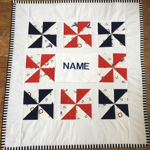 May include: A nautical-themed quilt with a white background and a black and white striped border. The quilt features pinwheel blocks in navy blue and red, with anchor and sailboat patterns. The center block has the word "NAME" in navy blue.