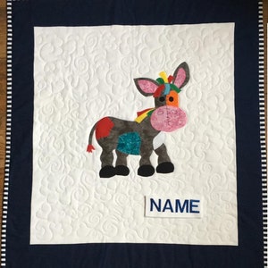 May include: A white quilt with a blue border and a patchwork donkey design. The donkey is made of different colored fabrics and has a pink nose. The quilt has a white label with the word "NAME" embroidered on it.