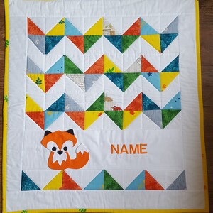 May include: A white baby quilt with a yellow border. The quilt features a chevron pattern of colorful triangles. A fox applique is in the center of the quilt with the word "NAME" embroidered below it.