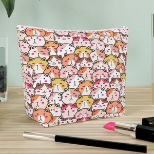 Op de afbeelding: Een roze en witte ritszak met een herhalend patroon van cartoonkatten in verschillende kleuren. De zak ligt op een houten oppervlak met een glazen vaas, een zwart-wit object en make-upkwasten.