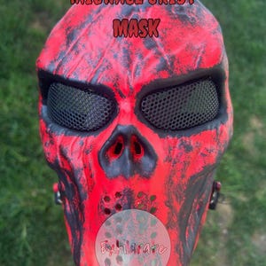 May include: A full-face skull mask featuring a vibrant red base with distressed black paint detailing. The mask includes black mesh eye coverings and a perforated nose and mouth area. The text MICHAEL CRIST MASK and Exhilarate are visible.