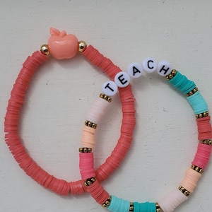 May include: Two handmade bracelets. One is coral with a peach-colored apple charm and gold beads. The other bracelet has the word "TEACH" spelled out in white letter beads, and is made of colorful beads in shades of pink, peach, teal, and white, with gold accents.