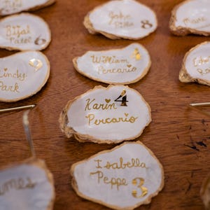 May include: White oyster shell place cards with gold lettering. Names and numbers are written on the shells. Some shells have gold leaf accents. The shells are arranged on a wooden surface, ideal for weddings or events.