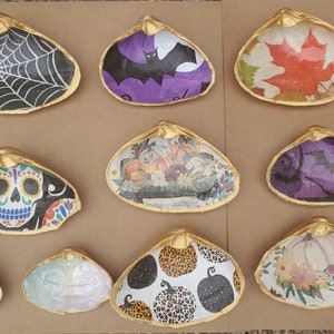 May include: A collection of decorative seashells with gold-painted rims. The shells feature various Halloween and autumn-themed designs, including a spiderweb, bat, maple leaf, sugar skull, pumpkins, and floral patterns. These shells are ideal for seasonal decor.
