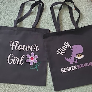 May include: Two black tote bags with white text and graphics. One says "Flower Girl" with a purple flower. The other says "Ring Bearer saurus" with a purple dinosaur holding rings.