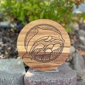 Puede incluir: Un posavasos redondo de madera con un diseño de un delfín saltando sobre las olas grabado con láser. El posavasos está hecho de madera marrón claro con un borde de madera marrón oscuro.