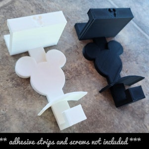 May include: Two 3D printed phone stands in the shape of Mickey Mouse heads. One is white and the other is black.  *** adhesive strips and screws not included ***
