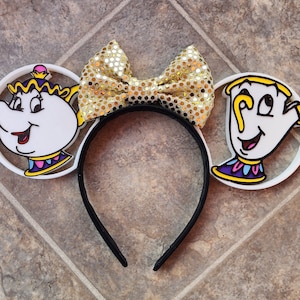 May include: White plastic headband with a gold glitter bow and two white plastic ears. One ear features a cartoon image of Mrs. Potts from Beauty and the Beast, and the other ear features a cartoon image of Chip from Beauty and the Beast.