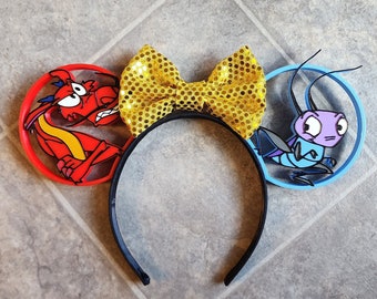 May include: A pair of black mouse ears with a yellow bow. The left ear features a red dragon with a yellow and red body. The right ear features a blue bug with a purple body.
