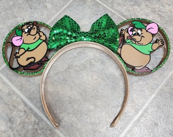May include: Green glitter bow headband with brown mouse ears featuring cartoon characters from the Disney movie The Rescuers. The mouse on the left ear is wearing a green shirt and the mouse on the right ear is wearing a green shirt and a green hat.