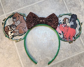 May include: Green headband with a brown glitter bow. The headband has two attached ears, one with a brown gorilla and a small human figure, and the other with a red elephant, a human figure, and a black gorilla.