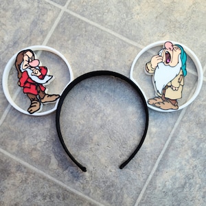 Puede incluir: Diadema de orejas de Mickey Mouse de plástico blanco con una diadema negra. Las orejas presentan una imagen de dibujos animados de dos enanos de la película de Disney Blancanieves y los Siete Enanitos. Un enano lleva un atuendo rojo y blanco y el otro lleva un atuendo azul y blanco.