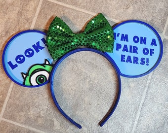 May include: Blue and green mouse ears headband with a green bow. The left ear has a blue background with the text "LOOK!" and a green monster eye. The right ear has a blue background with the text "I'M ON A PAIR OF EARS!"
