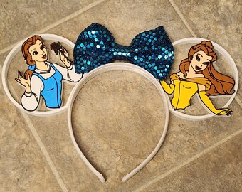 May include: White headband with two white ears featuring Belle from Beauty and the Beast. The ears have a blue sequined bow in the center.