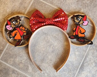 May include: A pair of brown mouse-themed Minnie Mouse ears with a red sequined bow. The ears feature a cartoon mouse wearing a red jacket and brown pants.