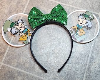 May include: A pair of white headband ears with a green sequin bow. The ears feature a cartoon image of the Mad Hatter from Alice in Wonderland.