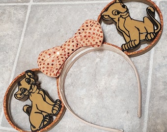 May include: A pair of brown and orange mouse ears with a sparkly orange bow. The ears feature a cartoon image of a lion cub.