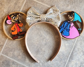 May include: A pair of brown mouse-themed Minnie Mouse ears with a silver glitter bow. The ears feature a cartoon image of a mouse wearing a red jacket and a mouse wearing a pink dress and blue jacket.