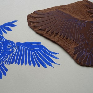 May include: A blue ink print of an owl with its wings spread out. The owl is facing to the left. The print is made from a woodblock carving of the owl, which is shown in the image.
