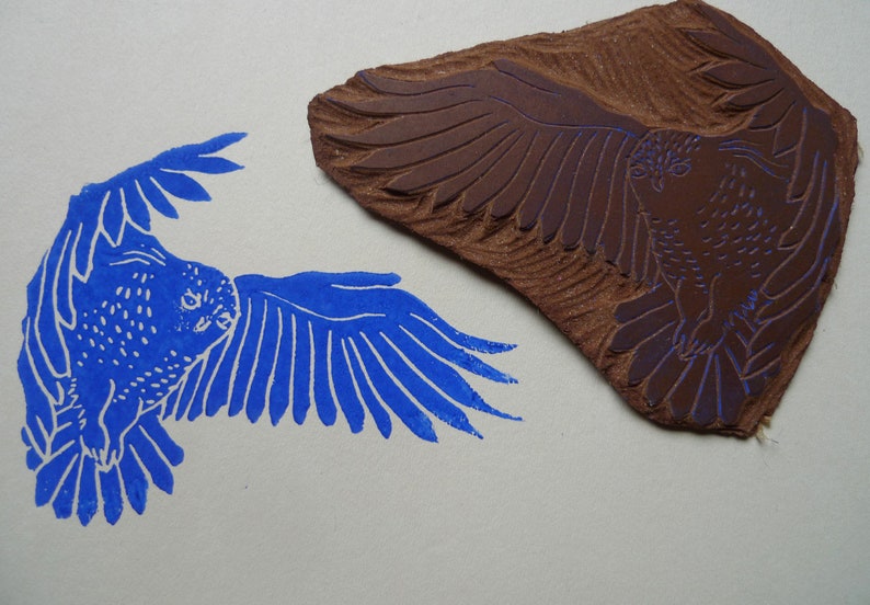 May include: A blue ink print of an owl with its wings spread out. The owl is facing to the left. The print is made from a woodblock carving of the owl, which is shown in the image.