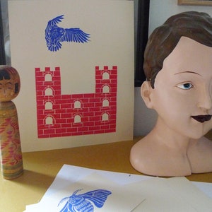 May include: A wooden doll with a painted face and colorful patterned clothing stands next to a print of a red brick castle and a blue bird in flight. The print is on a white background.