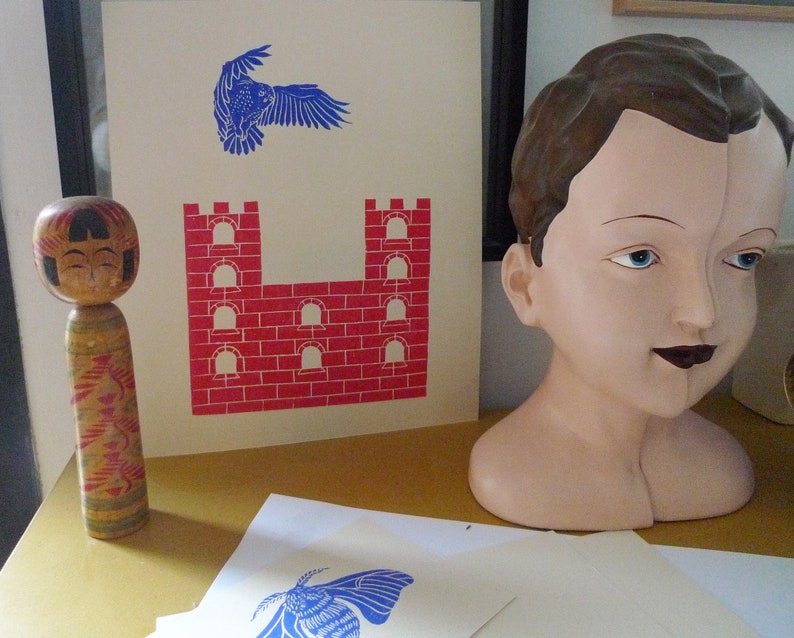 May include: A wooden doll with a painted face and colorful patterned clothing stands next to a print of a red brick castle and a blue bird in flight. The print is on a white background.