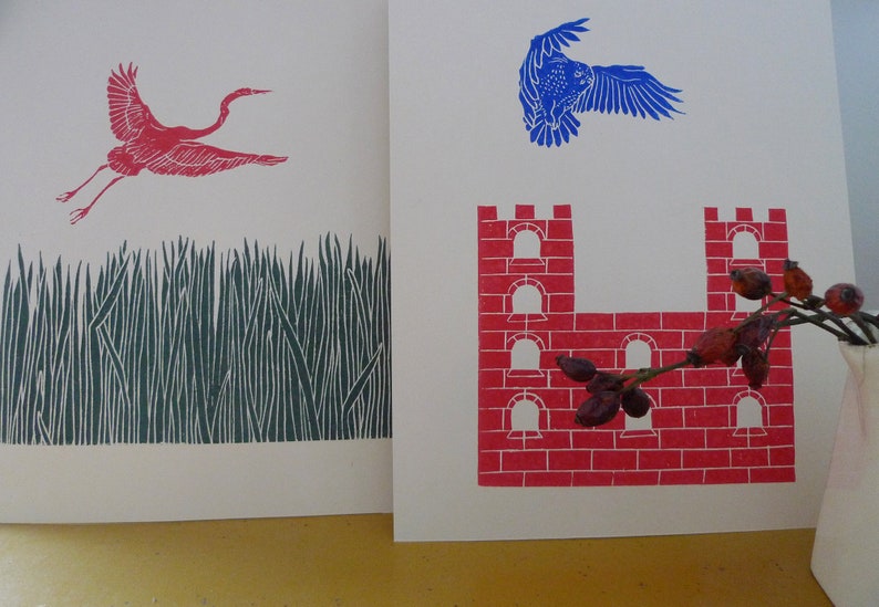 May include: Two pieces of art, one with a red bird flying over a green field and the other with a red brick castle with a branch of red berries.