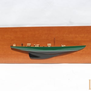 Könnte beinhalten: Ein Holzmodell der Yacht Shamrock V, ein grünes und schwarzes Segelschiff, montiert auf einer Holzplatte. Die Platte trägt ein Messing-Schild mit dem Text "Shamrock 1930".