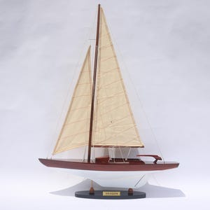 Könnte beinhalten: Ein Holzmodell-Segelboot mit rotem Rumpf und beigefarbenen Segeln. Das Modell trägt die Aufschrift "Dragon" und steht auf einem schwarzen Sockel. Die Segel sind gehisst, vor weißem Hintergrund.