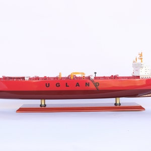 Puede incluir: Un modelo de barco rojo y blanco con el nombre "UGLAND" impreso en el lateral. El modelo está sobre una base de madera y es una réplica detallada de un barco real.