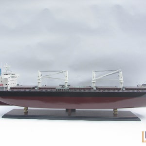 Pode incluir: Um modelo detalhado de um grande navio de carga com casco vermelho escuro, superestrutura branca e duas gruas brancas. O navio está sobre uma base de madeira.