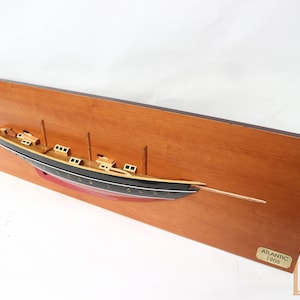 Könnte beinhalten: Ein hölzernes Schiffsmodell der Atlantic, gebaut im Jahr 1905, montiert auf einer Holzplatte. Das Schiff ist schwarz mit goldenen Akzenten und roten Details. Die Platte hat eine braune Holzmaserung und einen dunkelbraunen Rand. Die Platte trägt auch ein goldenes Etikett mit dem Text "ATLANTIC 1905" und ein Lux Ships Logo.