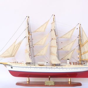 Puede incluir: Un modelo de madera detallado del barco de vela alemán Gorch Fock, con velas blancas y un casco rojo. El barco está sobre una base de madera y tiene una bandera alemana ondeando en el mástil.