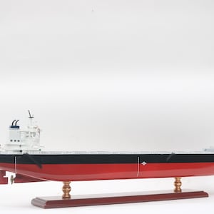 Pode incluir: Modelo de navio com casco vermelho, convés preto e superestrutura branca. O modelo é montado em uma base de madeira com suportes cor de latão. O navio tem detalhes, incluindo uma chaminé e antenas.