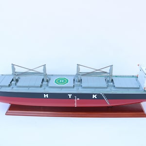 Pode incluir: Modelo detalhado de um navio de carga com casco vermelho, convés cinza e superestrutura branca. O navio tem as letras "HTK" na lateral e um heliporto verde e branco. Assenta numa base de madeira.