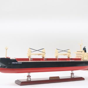 Pode incluir: Modelo do navio de carga "Crested Eagle". O navio tem um casco preto com fundo e convés vermelhos. Montado em uma base de madeira com placa de identificação. Possui dois guindastes e uma superestrutura.